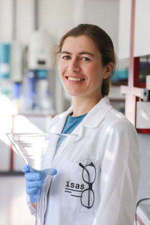 A Foto of Mona Ehlers in the laboratoy. She is wearing a lab coat and holding a glas funnel.