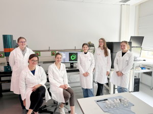 Das Bild zeigt einen Teil der Schülerinnen zusammen mit Luisa Röbisch, Dr. Anika Grüneboom und Dr. Christiane Stiller, während sie im Labor stehen und in die Kamera schauen.