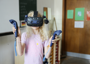 Marie steht im Klassenzimmer und trägt eine VR-Brille. In den Händen hält sie jeweils einen Controller.