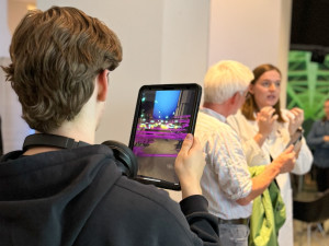 Ein Besucher hält ein Tablet in den Händen und betrachtet eine Projektion der Immunzellen. Im Hintergrund ist Lara Janz im Gespräch mit einem anderen Besucher.