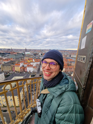 Felix Hormann stands on a viewing tower, camera around his neck. The sky and the roofs and buildings of a city can be seen in the background.