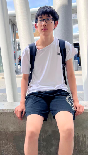 Lukas Fu, a 15-year-old with short black hair. He is wearing glasses and is sitting on a low wall.