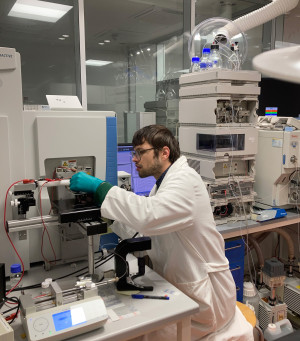Felix Hormann is wearing a white coat and sitting in a laboratory. He is positioning a sample under a mass spectrometer..
