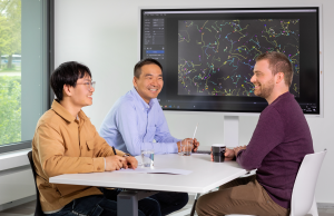 Das Bild zeigt Dr. Jianxu Chen mit den beiden Doktoranden Yu Zhou und Justin Sonneck an einem Tisch. Im Hintergrund ist ein Bildschirm mit einer ihrer Anwendungen zu sehen.
