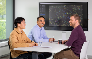 Das Bild zeigt Dr. Jianxu Chen mit den beiden Doktoranden Yu Zhou und Justin Sonneck an einem Tisch. Im Hintergrund ist ein Bildschirm mit einer ihrer Anwendungen zu sehen.