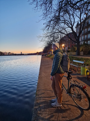 Felix Hormann steht mit seinem Fahrrad am Ufer eines Gewässers. Im Hintergrund ist der Sonnenuntergang sichtbar.