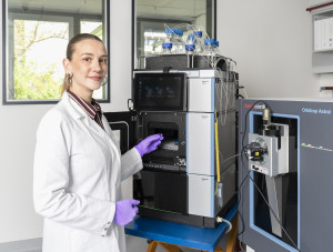 Neele Rottmann trägt einen weißen Laborkittel und violette Handschuhe und bedient in einem Labor ein Analysegerät zur Massenspektrometrie. Neben ihr befindet sich ein System zur Flüssigchromatographie, das über mehrere Schläuche mit einem Massenspektrometer verbunden ist. Auf dem Gerät stehen zahlreiche Probenbehälter. Während sie eine Probe in das System einsetzt, blickt sie in die Kamera.